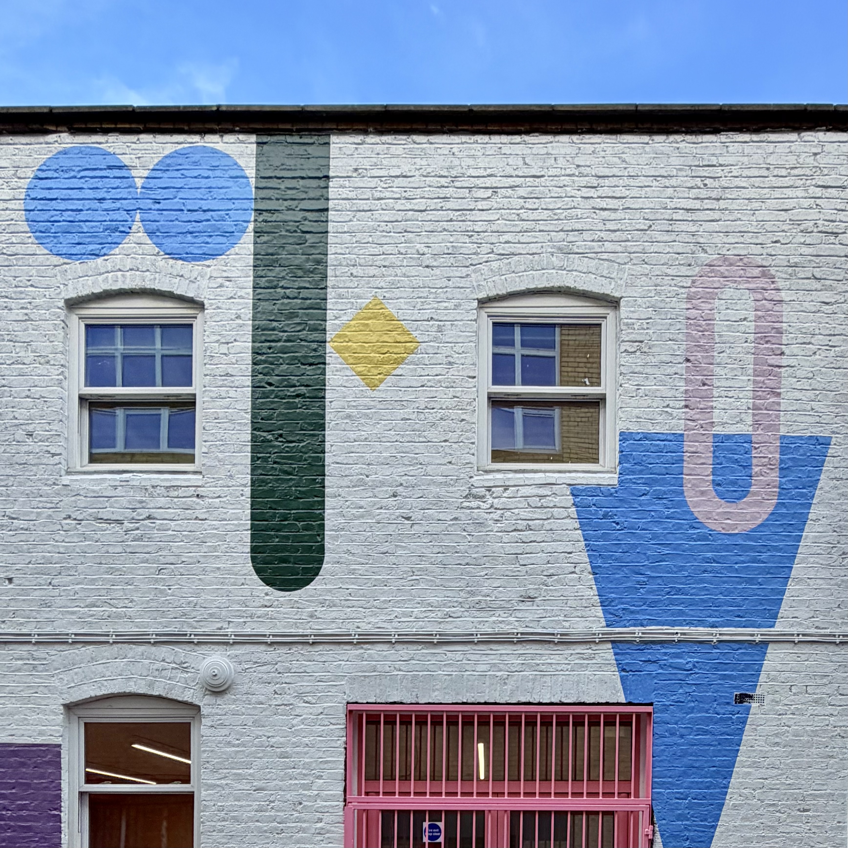 White brick building facade with colourful geometric mural shapes, two windows, and a pink gated doorway under a blue sky.