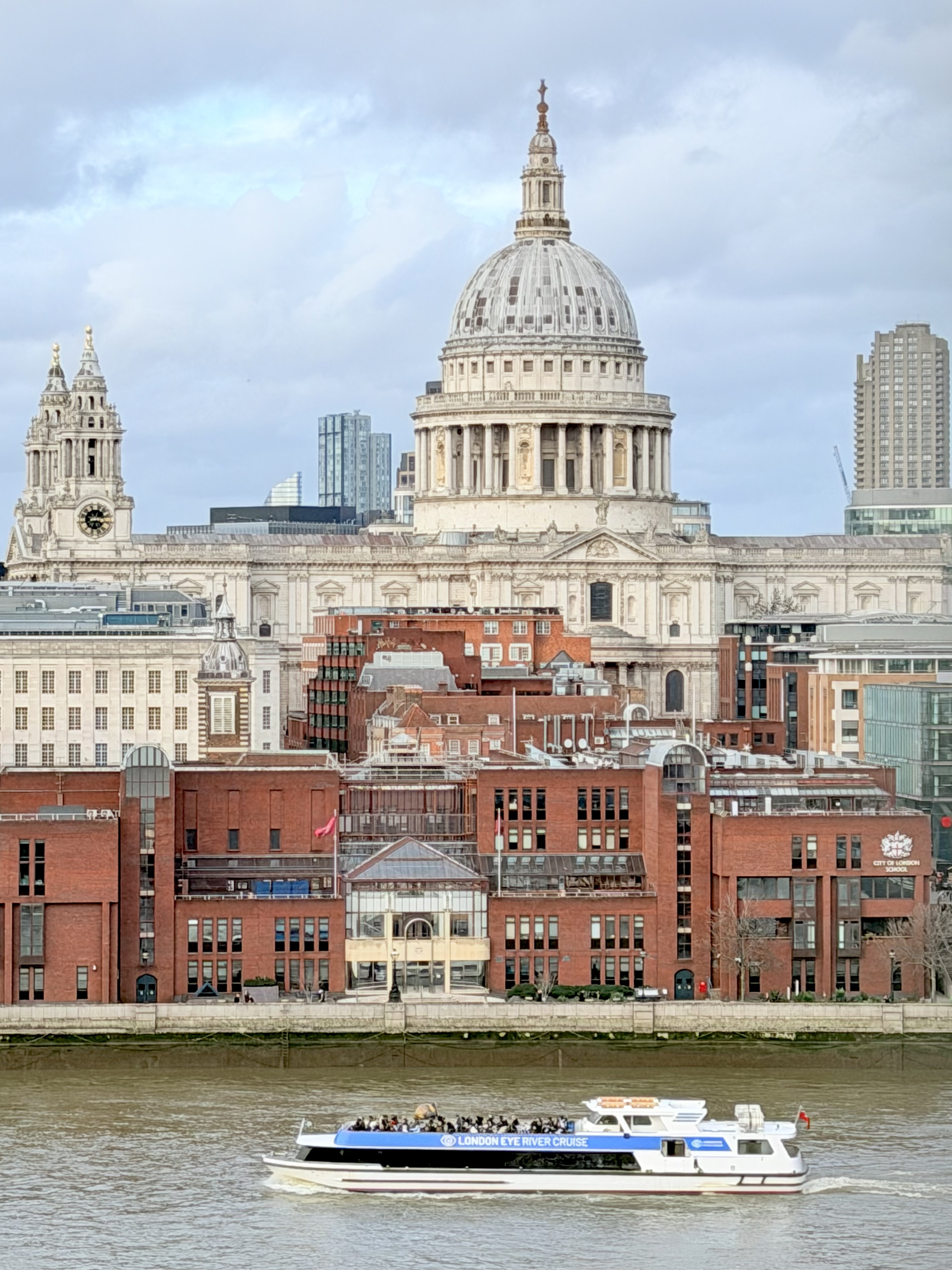 St Paul’s Cathedral rising above riverside buildings in London, with a sightseeing boat on the River Thames in the foreground under a cloudy sky.