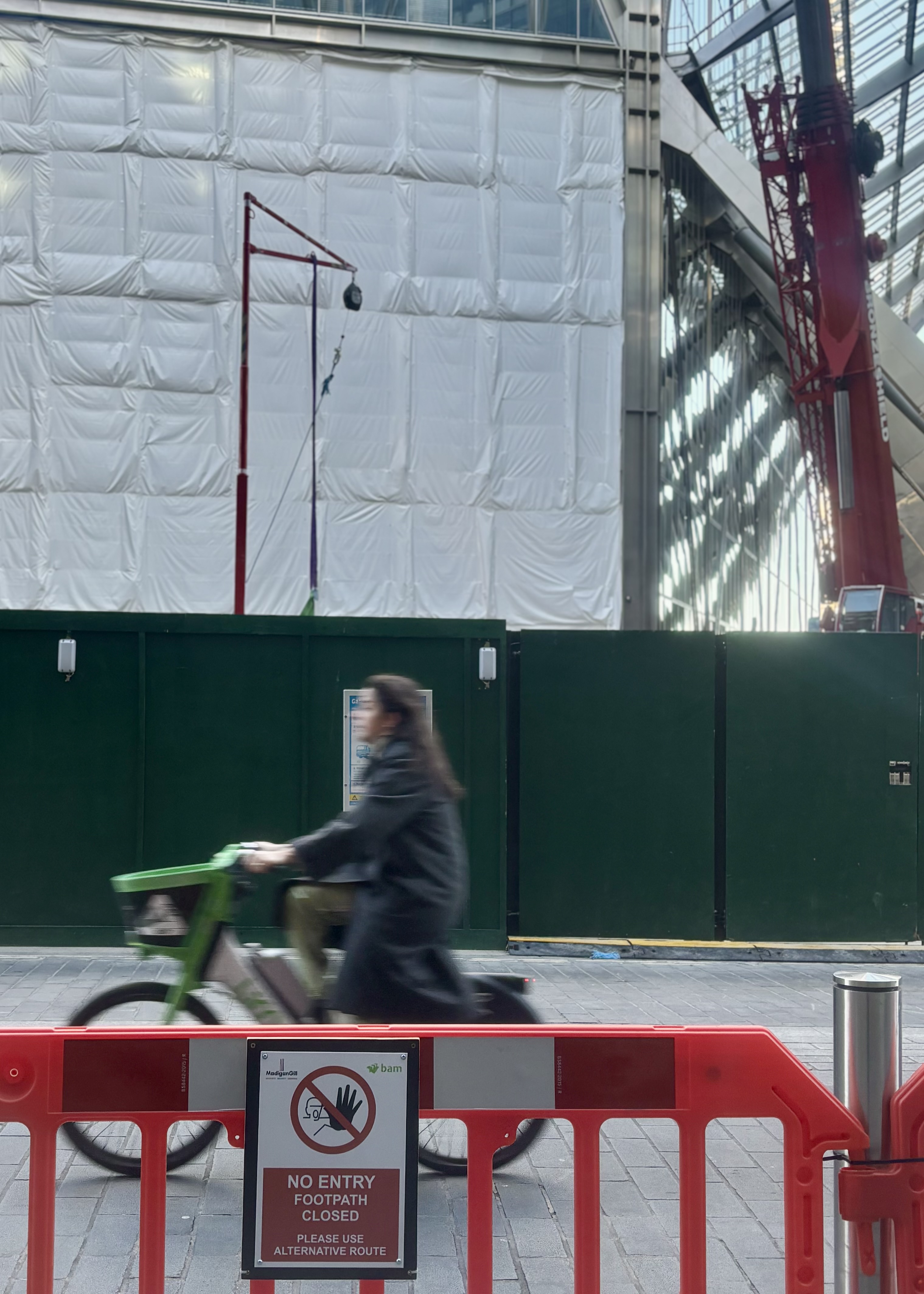 A cyclist rides past red barriers blocking a footpath, with a “No entry – footpath closed, please use alternative route” sign in the foreground and a large building under construction behind green fencing.