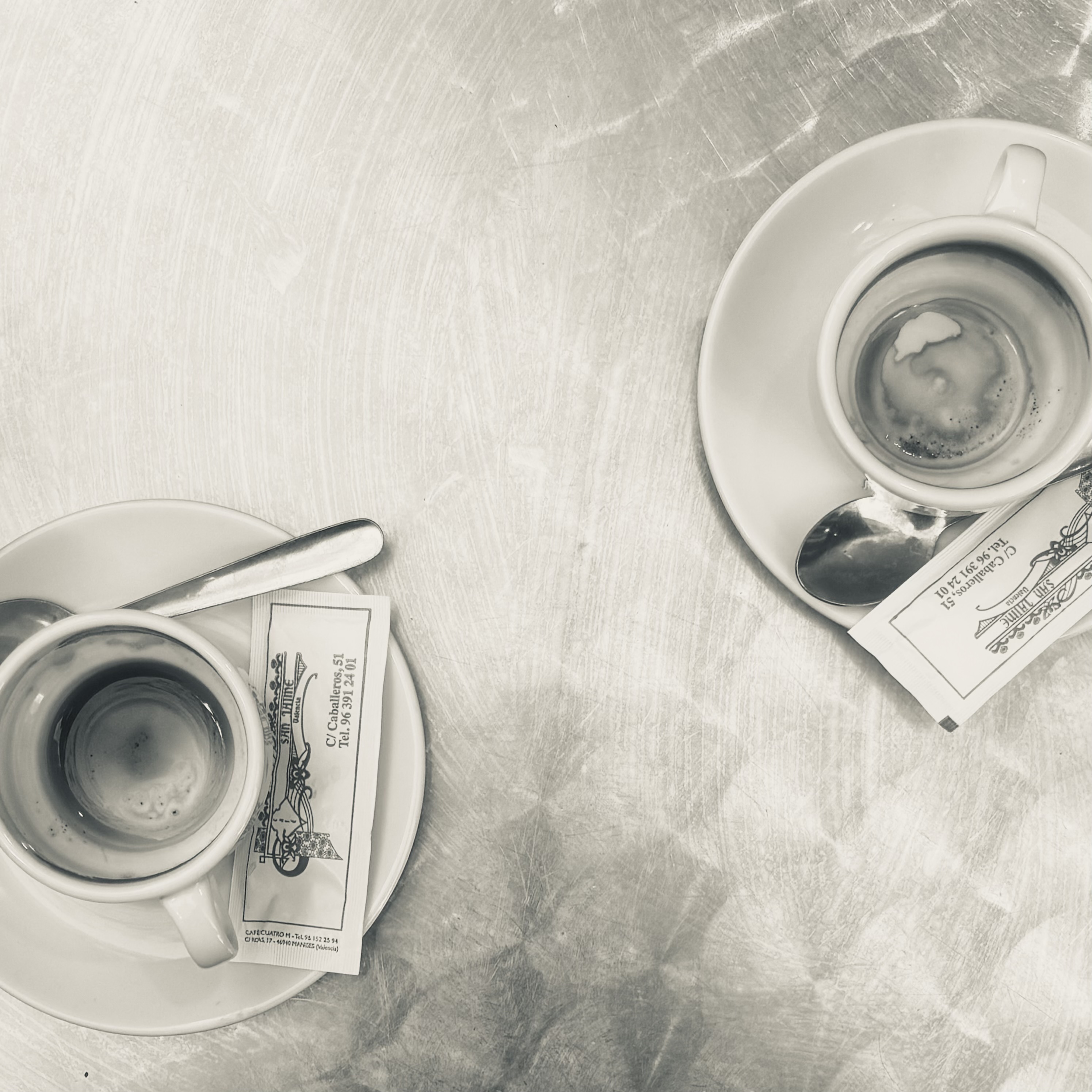 Overhead view of two espresso cups on saucers with sugar packets on a brushed metal table, positioned diagonally with space between them.