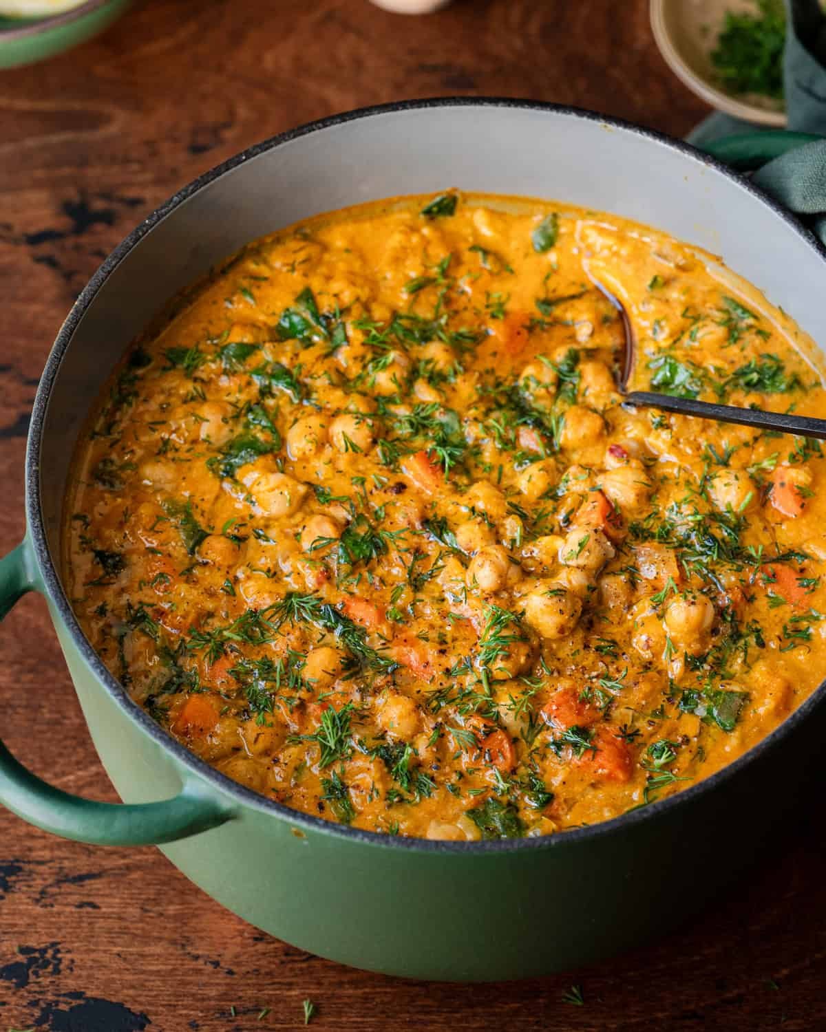 A large green pot filled with creamy chickpea and vegetable soup garnished with fresh dill, with a ladle resting inside.